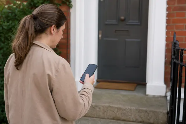 Personne consultant le suivi d'un colis en retard sur son téléphone devant sa porte d'entrée