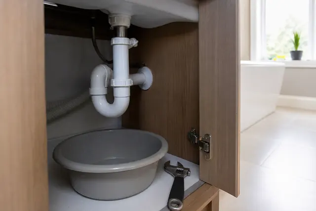 Siphon en plastique sous un lavabo de salle de bain, tuyauterie visible et clé de plombier posée à côté