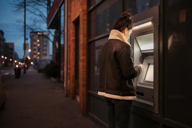 Personne face à un distributeur automatique de billets le soir, écran allumé, rue calme en arrière-plan
