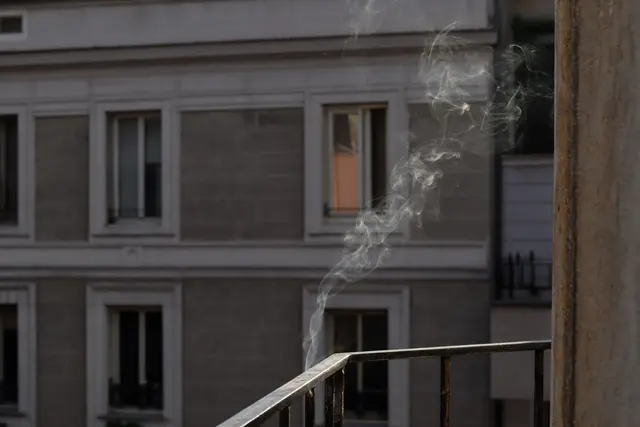 Fumée de cigarette s'élevant depuis un balcon d'immeuble résidentiel, vue rapprochée sur une façade avec fenêtres entrouvertes