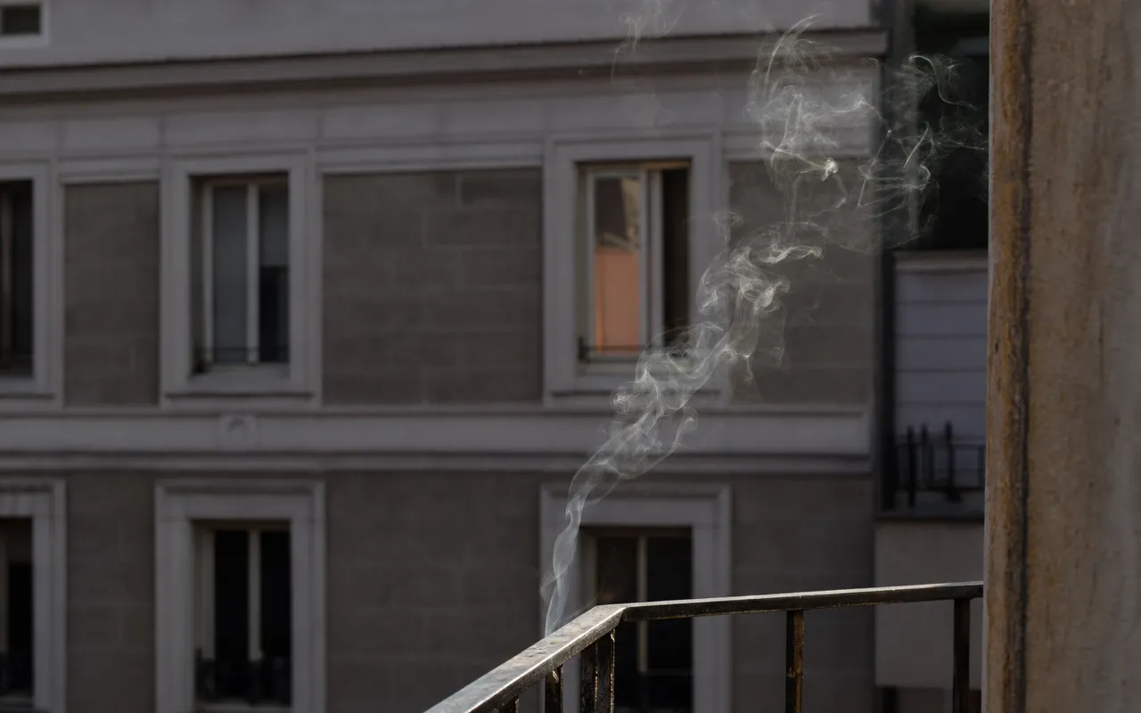 Fumée de cigarette s'élevant depuis un balcon d'immeuble résidentiel, vue rapprochée sur une façade avec fenêtres entrouvertes