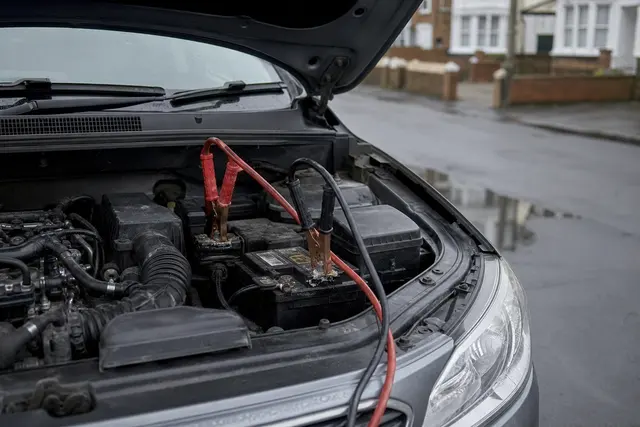 Capot de voiture ouvert dans une rue, câbles de démarrage rouge et noir pincés sur les bornes de la batterie